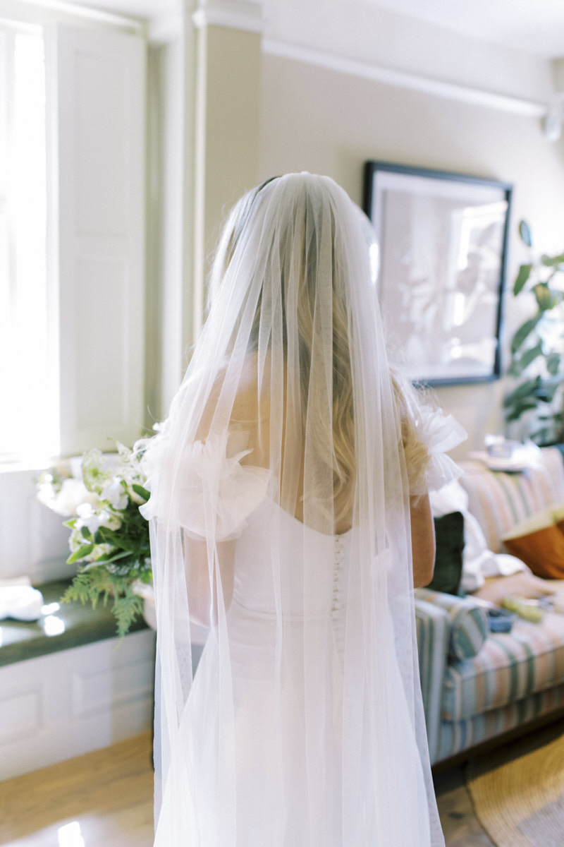 the bride looking outside the cottage window and wearing floor length veil