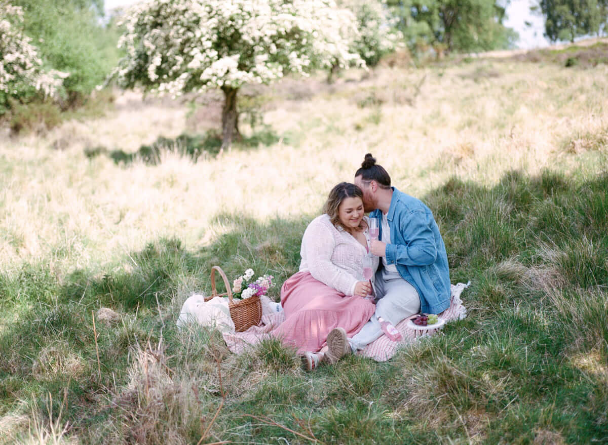 couple sitting on the picnic blanket in open field with picnic basket next to them