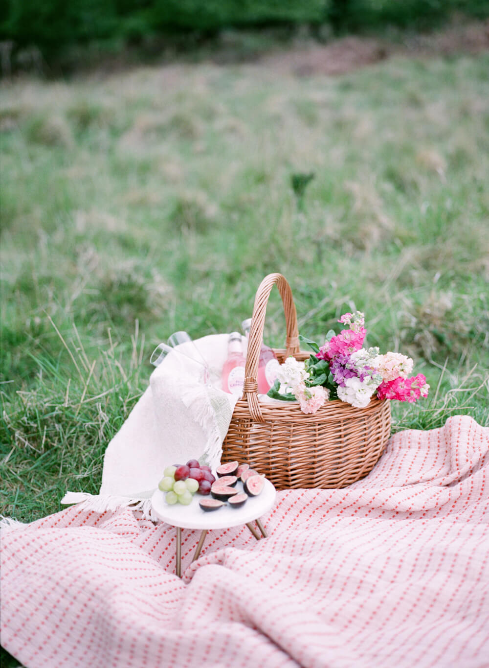 romantic outdoor picnic set up for pre-wedding photoshoot with pink cotton blanket, round wicker basket filled with flowers, lemonade and snacks like grapes and figs