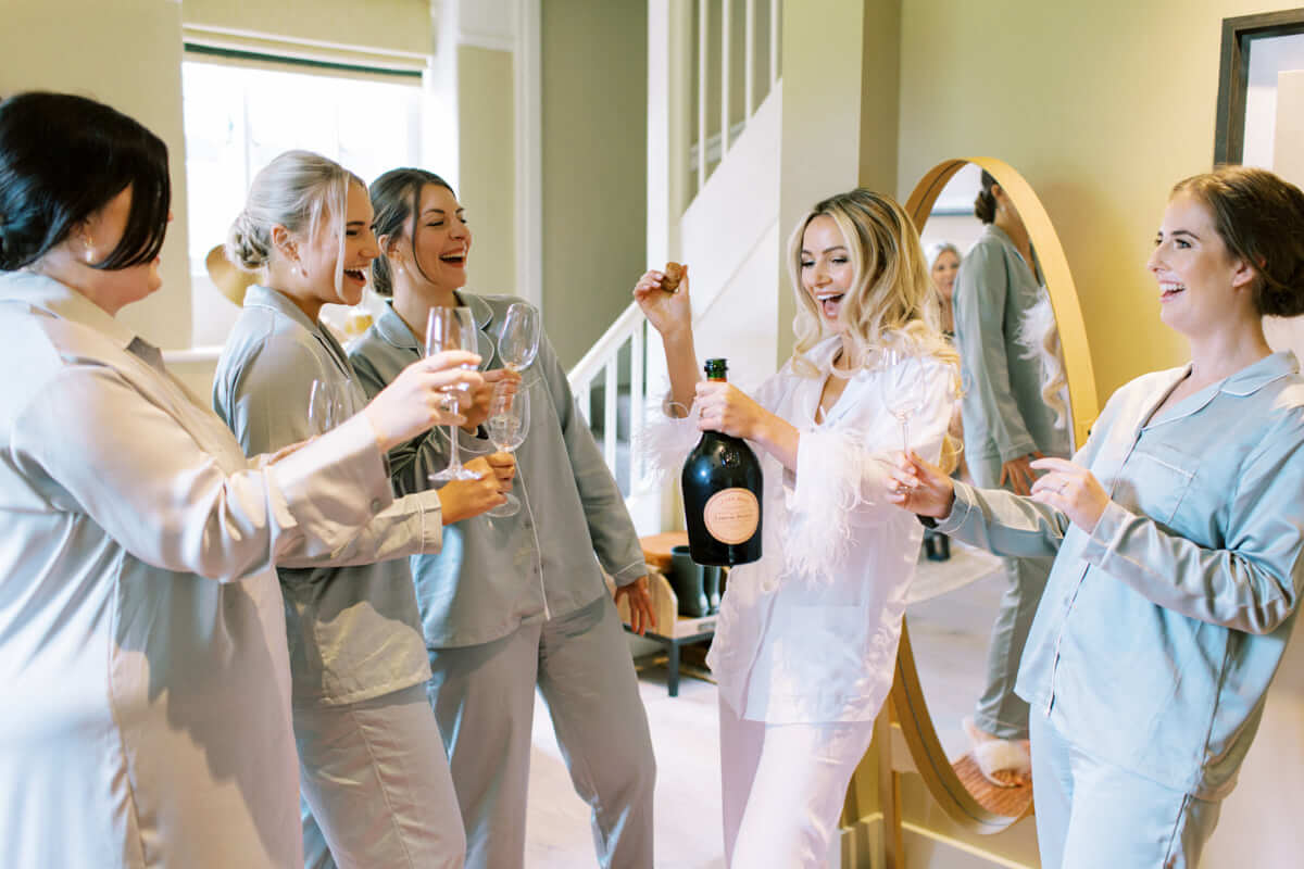 Bride and bridesmaids wearing satin dressing gowns opening a champagne bottle, showing a stress-free wedding schedule by laughing